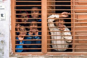 Group of children looking through the bars of a wooden window with curious expressions on their faces.